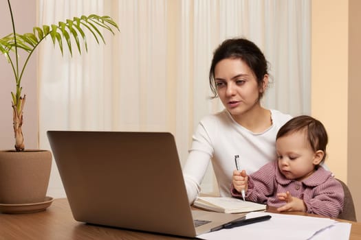 Cheerful pretty businesswoman working on laptop at home with her little child girl