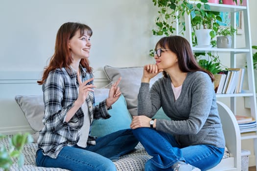 Talking happy smiling mother and teenage daughter sitting together on couch at home. Family, communication, motherhood, friendship, relationship between parent and daughter 18-20 years old