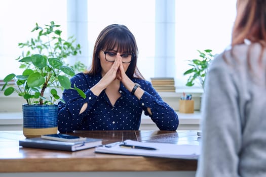 Mature sad woman in therapy session with mental professional psychologist, sitting at table in office of therapist counselor social worker. Psychology psychotherapy mental assistance treatment support