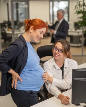 Caucasian woman touching belly of pregnant colleague while talking in office