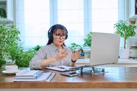 Mature woman in headphones having video conference using laptop computer in home office. Remote virtual meeting, online training, consulting, testing. Work, education, technology, people concept