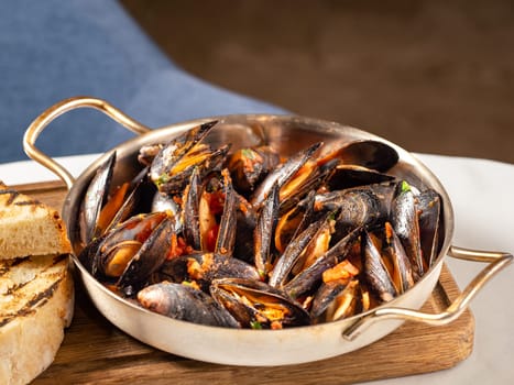 Delicious black mussels in red tomato sauce. Mediterranean black mussels in metal pan on wooden cutting board with grilled bread on restaurant table background