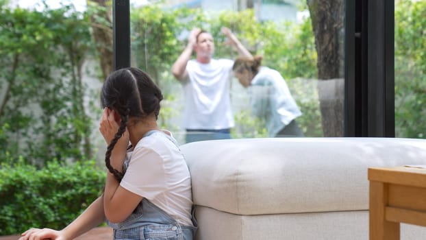 Stressed and unhappy young girl huddle in corner, cover her ears blocking sound of her parent arguing in background. Domestic violence at home and traumatic childhood develop to depression. Synchronos