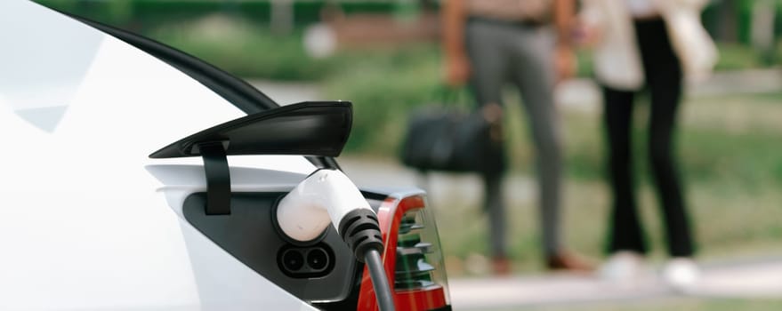 Young couple recharge electric car battery from charging station in green city park in springtime. Rechargeable EV car for sustainable environmental friendly urban travel lifestyle. Panorama Expedient