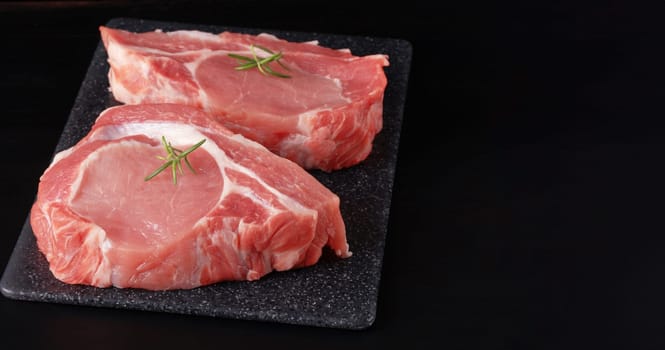 Raw pork steak with rosemary on a black table.