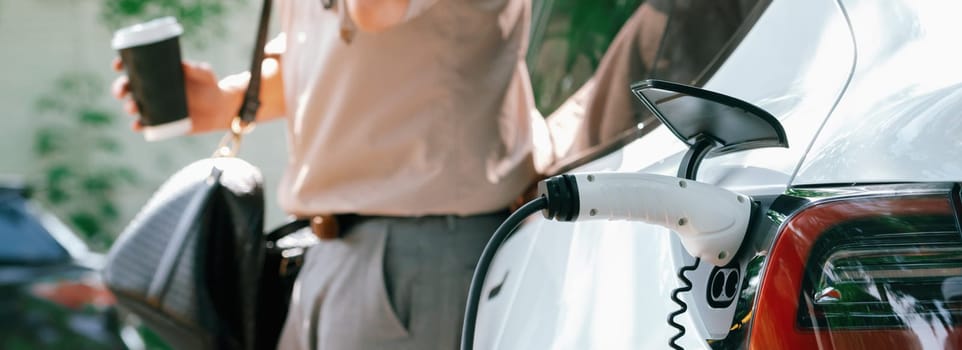 Young man with coffee cup, recharge electric car's battery from EV charging station in city commercial parking lot. Eco friendly EV car with urban and shopping lifestyle. Panorama Expedient