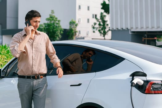 Young man recharge EV electric vehicle at green city commercial mall parking lot while talking on phone. Sustainable urban lifestyle for eco friendly EV car with battery charging station. Expedient