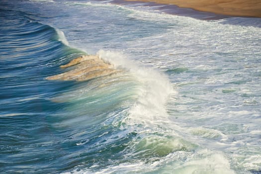 A closeup shot of the ocean with wind waves crashing onto the shore, creating a beautiful pattern of furrows in the water. The landscape is enhanced by cumulus clouds in the sky