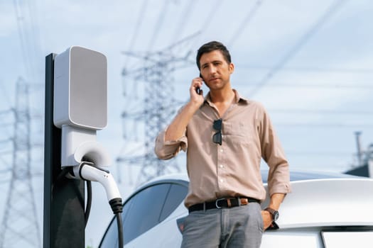 Man talking on the phone while recharge EV car battery at charging station connected to power grid tower electrical as electrical industry for eco friendly car utilization.Expedient