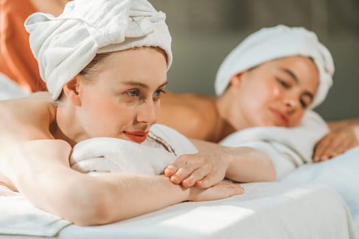 A couple of beautiful young woman with white towel lie on comfortable spa bed during receive relaxing body massage by professional masseur surrounded at spa salon. Gray background. Tranquility.