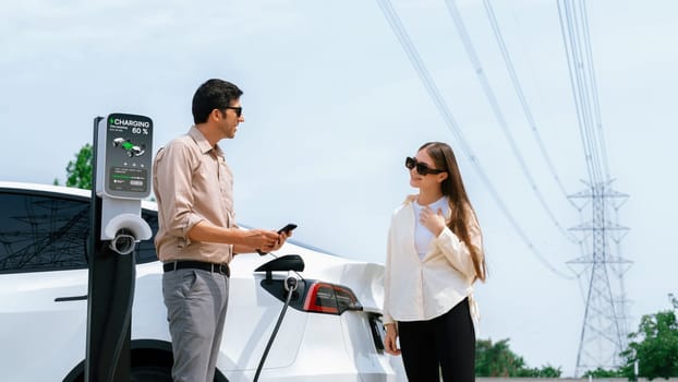 Couple pay for electricity with smartphone while recharge EV car battery at charging station connected to power grid tower electrical as electrical industry for eco friendly car utilization.Expedient