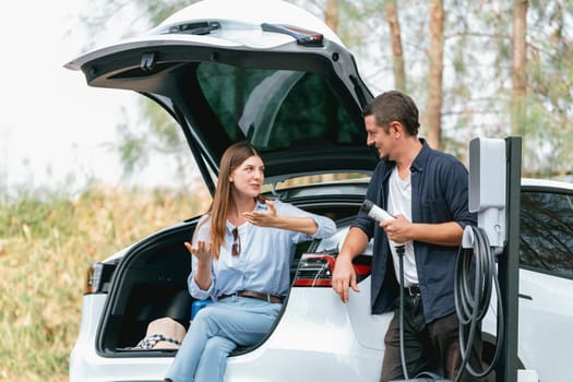 Lovely young couple recharging battery for electric car while relaxing and sitting on the trunk during road trip travel EV car in autumnal forest. Eco friendly travel on vacation during autumn. Exalt