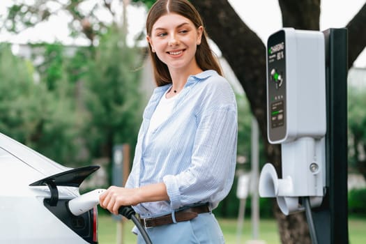 Young woman recharge EV electric vehicle's battery from EV charging station in outdoor green city park scenic. Eco friendly urban transport and commute with eco friendly EV car travel. Exalt