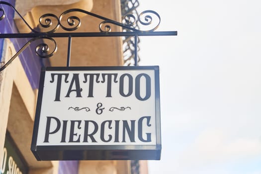 A sign with bold font hangs from a pole on the side of the building showcasing the tattoo and piercing shop, adding an edgy fixture to the facade