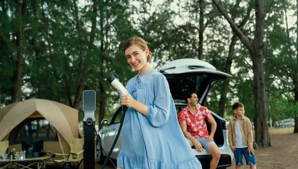 Outdoor adventure and family vacation camping at sea travel by eco friendly car. Cheerful woman or mother holding, pointing EV charger point with playful and happiness posture in campsite. Perpetual