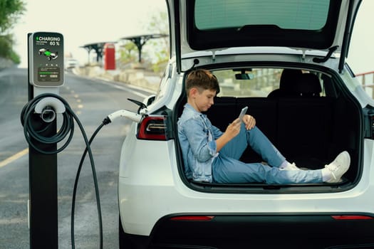 Little boy sitting on car trunk, using smartphone while recharging eco-friendly car from EV charging station. EV car road trip travel as alternative vehicle using sustainable energy concept. Perpetual