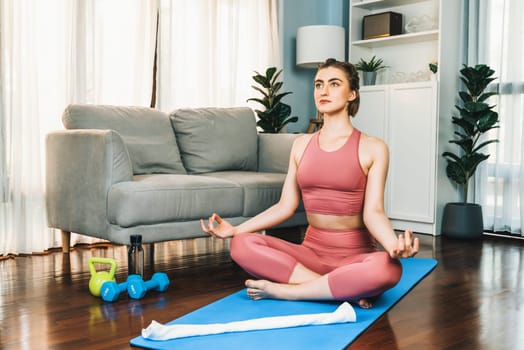 Flexible and dexterity woman in sportswear doing yoga position in meditation posture on exercising mat at home. Healthy gaiety home yoga lifestyle with peaceful mind and serenity.