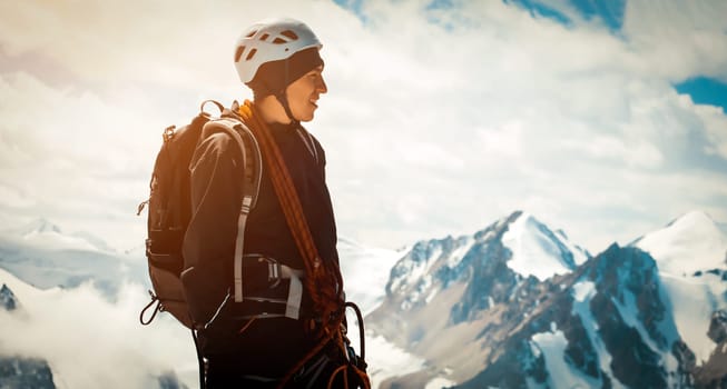 A young man traveler is engaged in mountaineering. In a helmet, with a rope, a harness, gloves, climbs to the top, against the backdrop of a stunning view with snow-capped mountains.