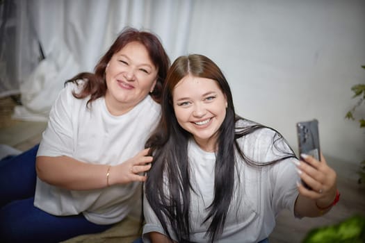 Fat funny funny adult mother and daughter posing, taking selfies indoors. Body positive, friendly family in the room