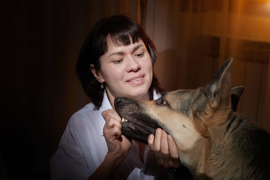 Adult mature woman with big shepherd dog in white shirt in dark room. Concept of loneliness and love for animals, pets