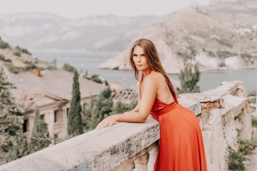 Side view a Young beautiful sensual woman in a red long dress posing on a volcanic rock high above the sea during sunset. Girl on the nature on overcast sky background. Fashion photo