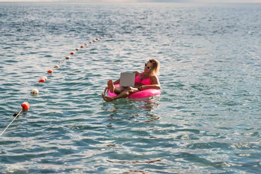 Woman works on laptop in sea. Freelancer, blond woman in sunglases floating on an inflatable big pink donut with a laptop in the sea at sunset. People summer vacation rest lifestyle concept