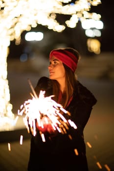 Woman holding sparkler night while celebrating Christmas outside. Dressed in a fur coat and a red headband. Blurred christmas decorations in the background. Selective focus.