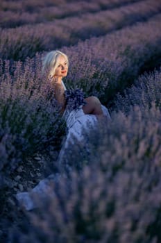 Blonde woman poses in lavender field at sunset. Happy woman in white dress holds lavender bouquet. Aromatherapy concept, lavender oil, photo session in lavender.