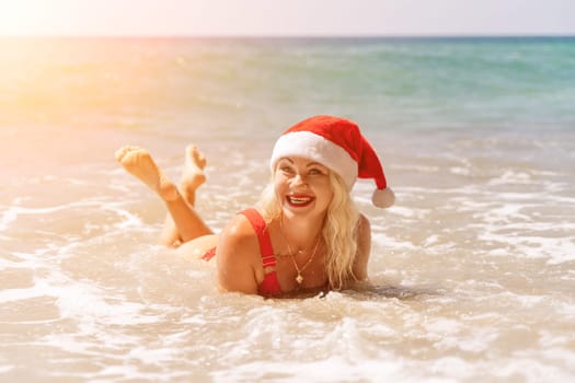 Female beach Santa hat wave coast. beach relaxation seaside. A woman in a red swimsuit enjoying her time on the beach, lying on the sand and being covered by a wave