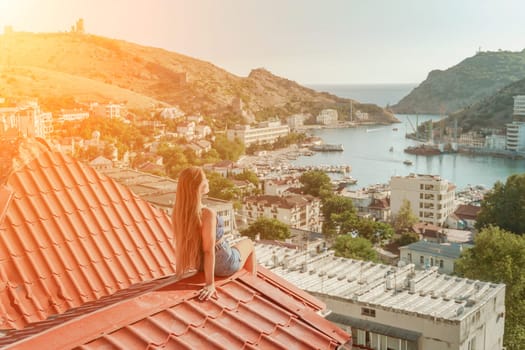 Woman sits on rooftop, enjoys town view and sea mountains. Peaceful rooftop relaxation. Below her, there is a town with several boats visible in the water. Rooftop vantage point