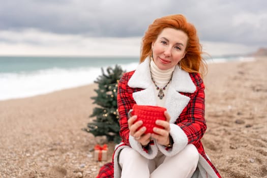 Lady in plaid shirt with a red mug in her hands enjoys beach with Christmas tree. Coastal area. Christmas, New Year holidays concep.