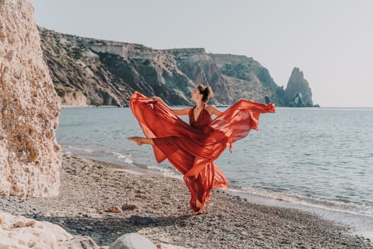 Woman red dress sea. Female dancer in a long red dress posing on a beach with rocks on sunny day. Girl on the nature on blue sky background