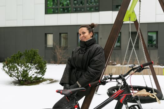 Lifestyle concept, a young brunette woman in a black winter jacket walks near the house with a bicycle.