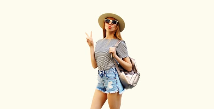 Portrait of young woman blowing her lips sending kiss wearing summer straw hat, backpack and shorts on white background