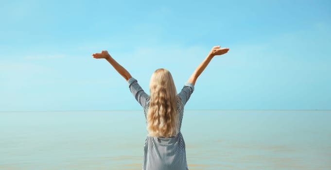 Summer vacation, happy young woman raising her hands up on the beach on sea coast and blue sky background