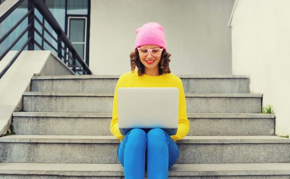 Portrait of stylish modern young woman working with laptop wearing a colorful clothes, eyeglasses in the city