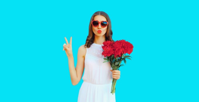 Portrait of beautiful woman with bouquet of red rose flowers and blowing her lips sending kiss in heart shaped sunglasses on blue background
