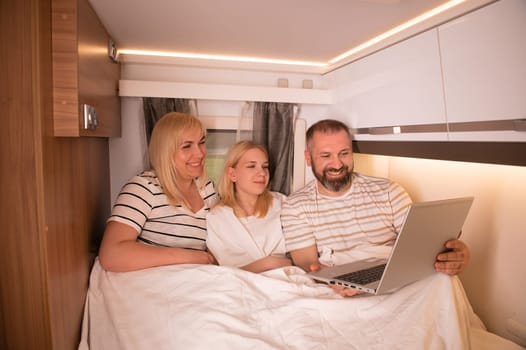 A family of three is watching a movie on a laptop while sitting in the bed of their motorhome.