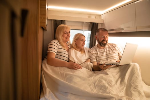 A family of three is watching a movie on a laptop while sitting in the bed of their motorhome.