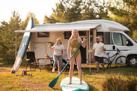 the family is resting next to their mobile home. My daughter is standing with a paddle on a sup board, and her parents pour water on her.