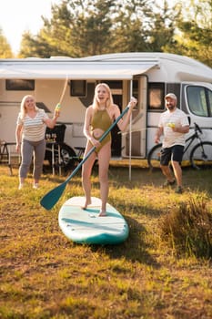 the family is resting next to their mobile home. My daughter is standing with a paddle on a sup board, and her parents pour water on her.