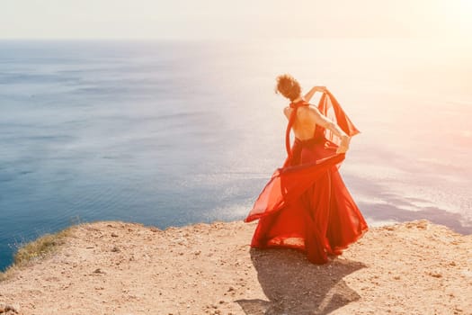 Side view a Young beautiful sensual woman in a red long dress posing on a rock high above the sea during sunrise. Girl on the nature on blue sky background. Fashion photo.