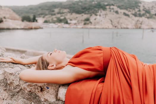 Side view a Young beautiful sensual woman in a red long dress posing on a volcanic rock high above the sea during sunset. Girl on the nature on overcast sky background. Fashion photo