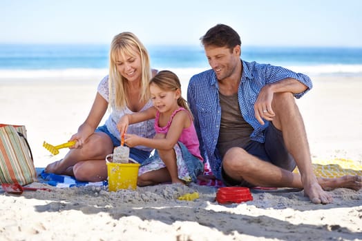 Mom, dad and girl with sandcastle by sea on vacation with care, learning or building on holiday in summer. Father, mother and daughter with sand in plastic bucket for game, blanket and happy at beach.