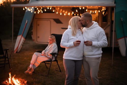 A married couple with glasses of wine stands against the background of a motorhome and rests together by the campfire. Evening family vacation.