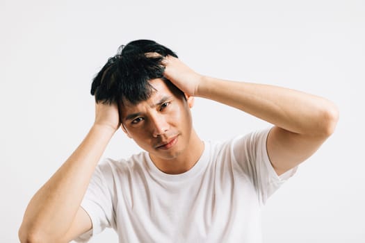 Portrait of a young Asian man, looking sad and exhausted, holding his head in despair due to stress and a headache. Studio shot isolated on white, depicting suffering and depression.