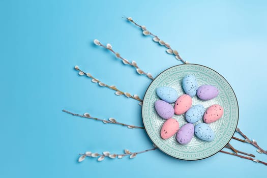 Dish with colored Easter eggs and willow branches with catkins on the blue background. Top view.