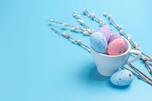 Porcelain cup with colored Easter eggs and willow branches with catkins on the blue background. Top view.