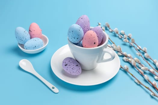 Porcelain cup with colored Easter eggs on a saucer, a spoon and willow branches with catkins on the blue background. Top view.