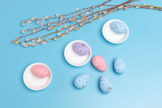 Porcelain saucers with colored Easter eggs and willow branches with catkins on the blue background. Top view.